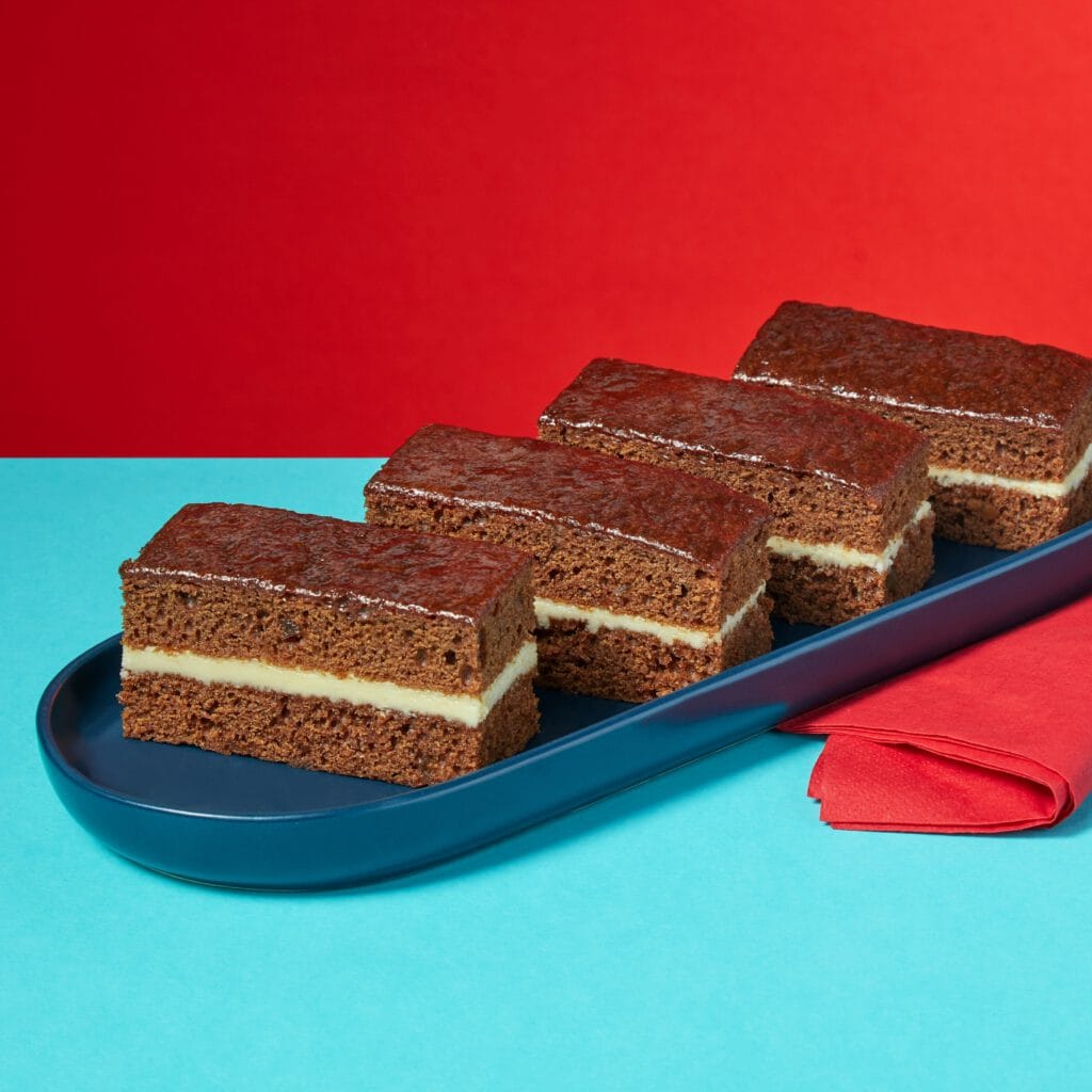 Four slices of Rum and Ginger Traycake on a dark blue oblong plate sitting on a red napkin on red and light blue background.