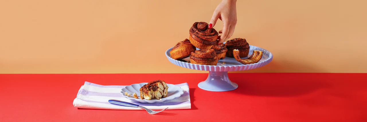 Fresh Cake Club cinnamon buns stacked on a stand with a hand reaching in and a sliced cinnamon bun on a plate.