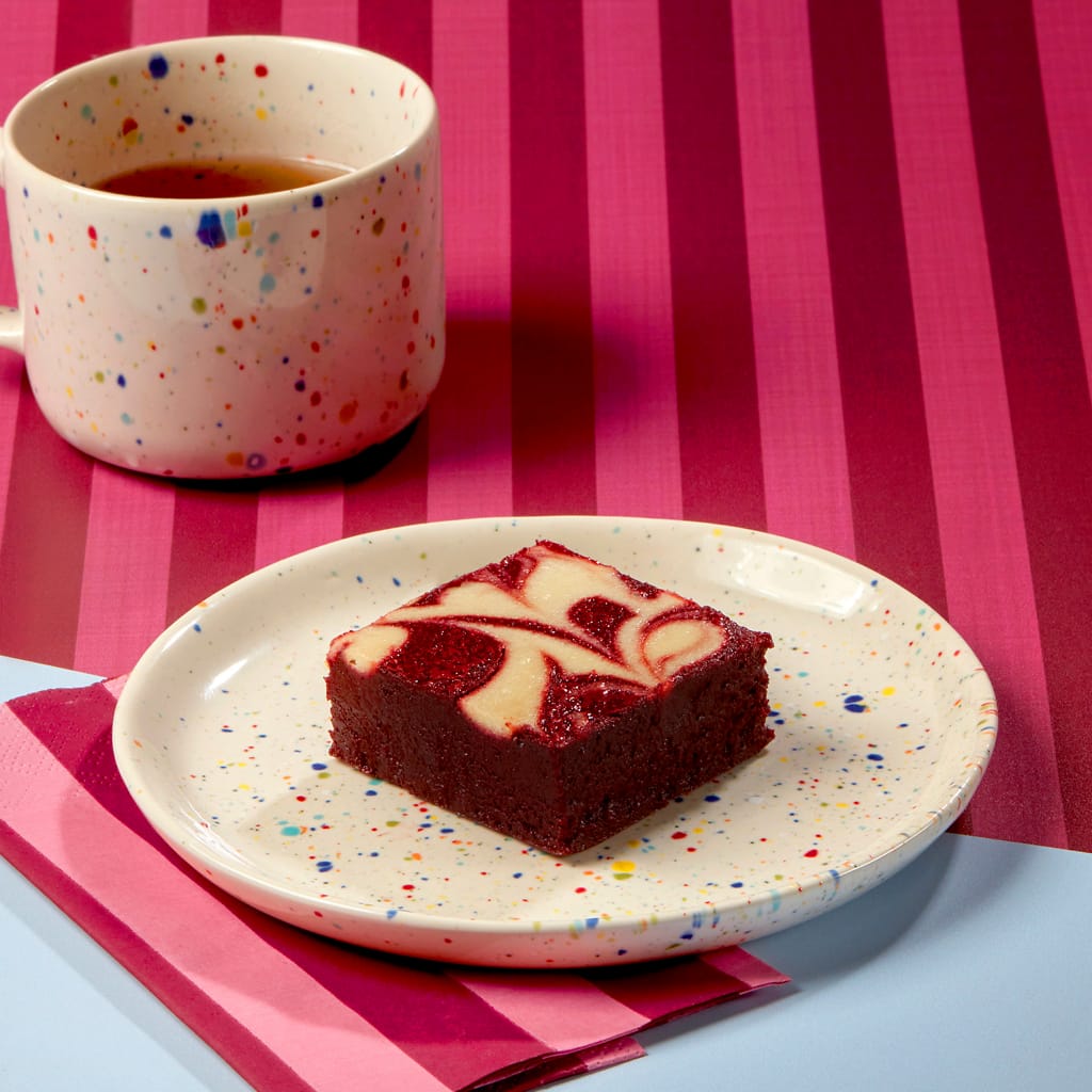 A succulent slice of Cake Club's Red Velvet Brownie on a spotted plate next to a spotty coffee cup on a claret and light blue background.