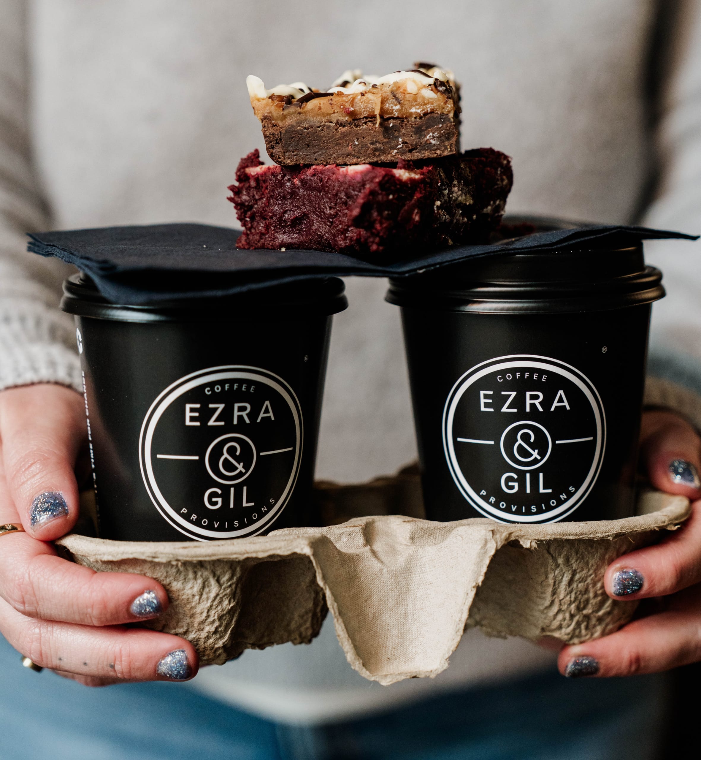 A slice of Red Velvet Cheesecake Brownie and Salted Caramel and Comb Brownie on top of two Ezra & Gil takeaway cups.