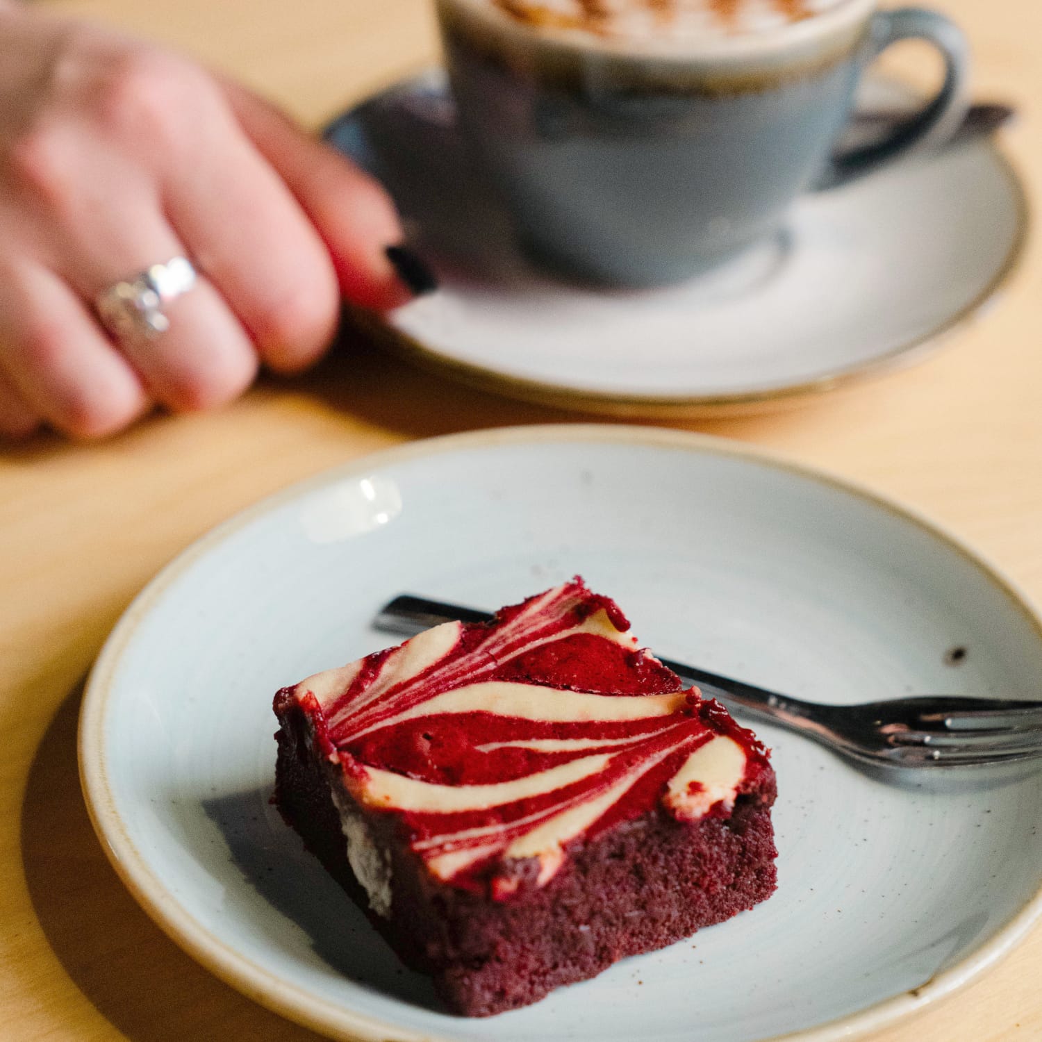 A slice of Cake Club's Red Velvet Cheesecake Brownie on a plate in a coffee shop next to a hand touching a coffee cup on a saucer.