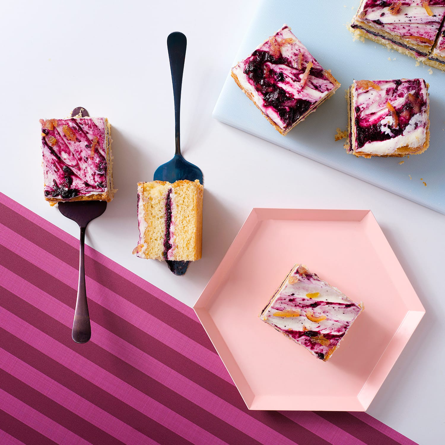 an overhead image of sliced lemon blueberry cake served with cake servers on white and pink stripy background