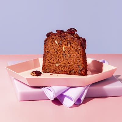 a slice of banana pecan and rye loaf cake on a pink board with a purple napkin and a pink plate on a light blue background